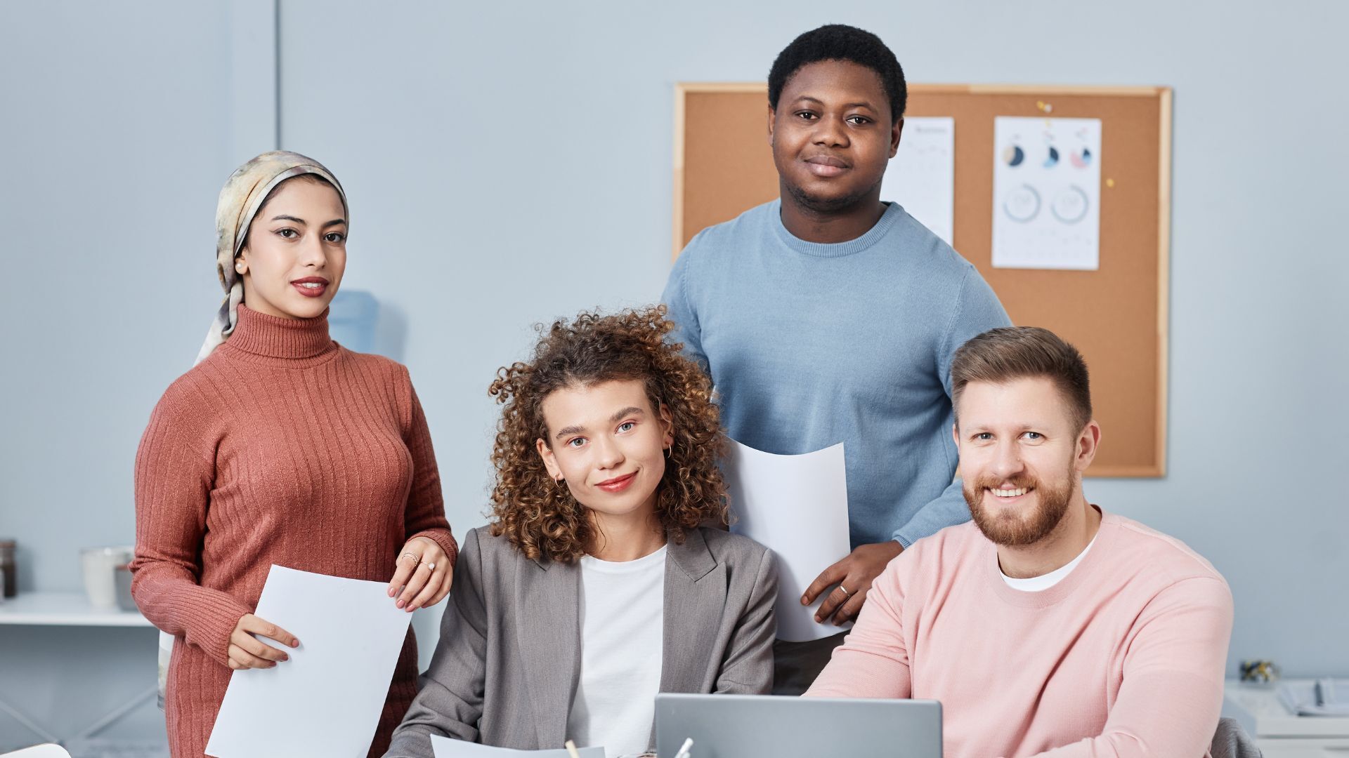 L'image montre un groupe diversifié de quatre collègues souriants dans un bureau moderne. Ils tiennent des documents et un ordinateur portable est visible, suggérant une ambiance de travail collaborative.